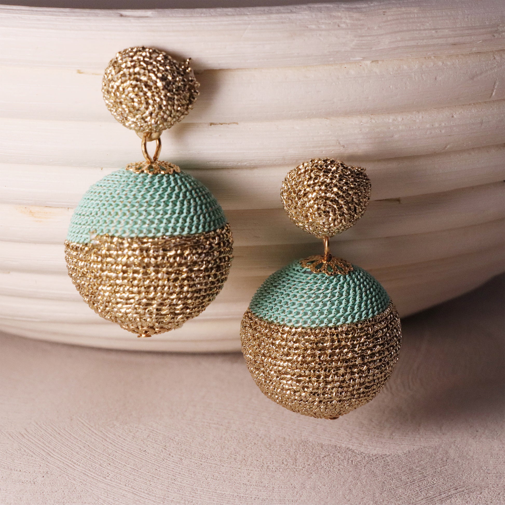Macro shot of two rattan straw spheres with natural grain texture, dangling from gold chains against a sunset-hued textile
